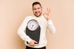 A happy male patient holding a weight scale and giving an "OK" hand gesture to celebrate reaching his weight loss goal.